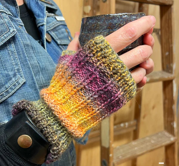 you see the torso of a person wearing a jean jacket and fingerless gloves drinking some tea. there's a plywood wall and a ladder in the background.