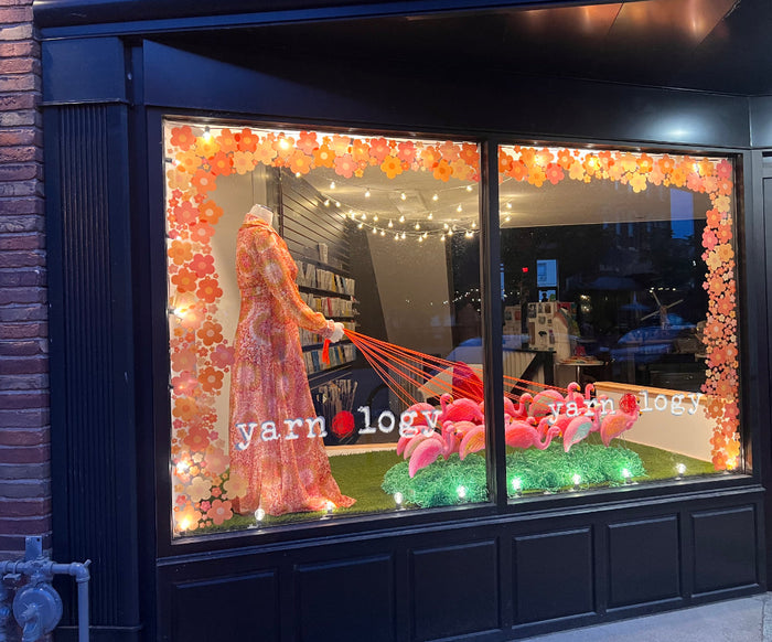 A look at Yarnology's front window at night. There's orange, gold, and salmon  flower decals running along both sides and the top. There's a mannequin in an old fashioned long dress with flowers of the same colors walking a group of pink flamingos.  