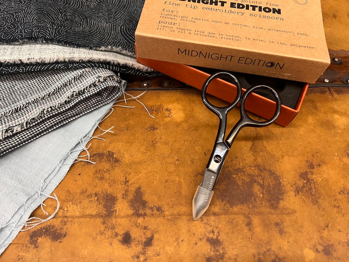 Scissors with their tip protector on, with their  box above them and a stack of fabric to the left.