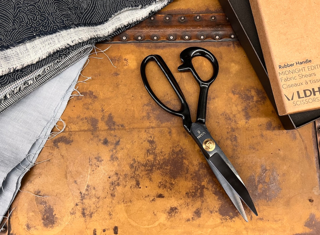 Scissors with their box to the right and a stack of fabric to the left.