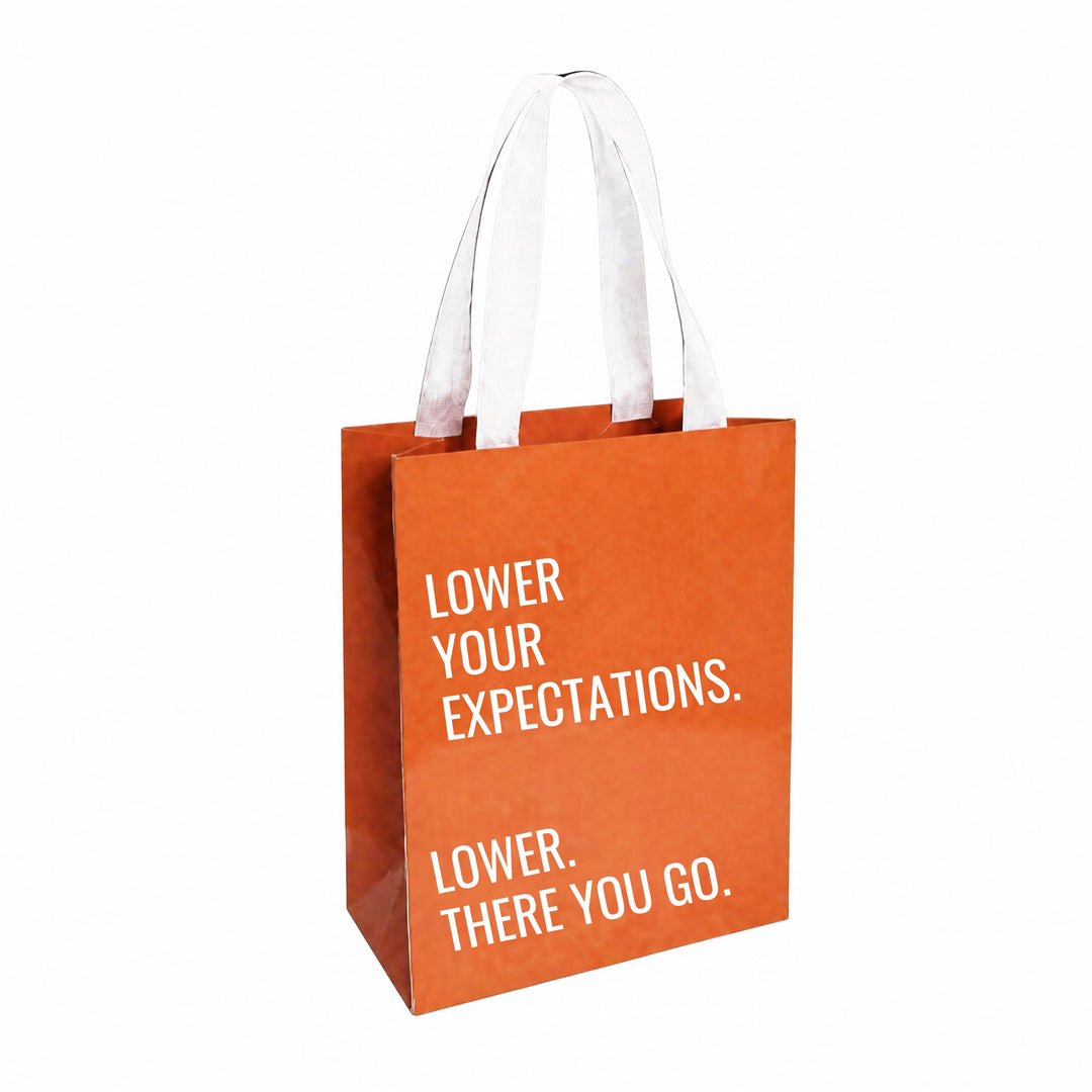 Orange gift bag with white handles. The bag reads, " LOWER YOUR EXPECTATIONS. LOWER. THERE YOU GO."