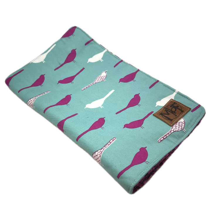 The outside of the needle case with a teal background and an all-over bird print in magenta and white. A brown leather patch with the brand name MbF is sewn onto the right edge.