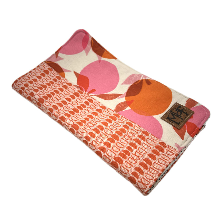 The cover of the knitting needle holder. One side is an abstract pattern that is reminiscent of citrus fruit in tangerine oranges and light pinks.  The other side features the same colors in rows of half moons. Alternating rows of outlined half-moons and filled in half moons. There's a leather patch in the lower corner with MbF on it. 