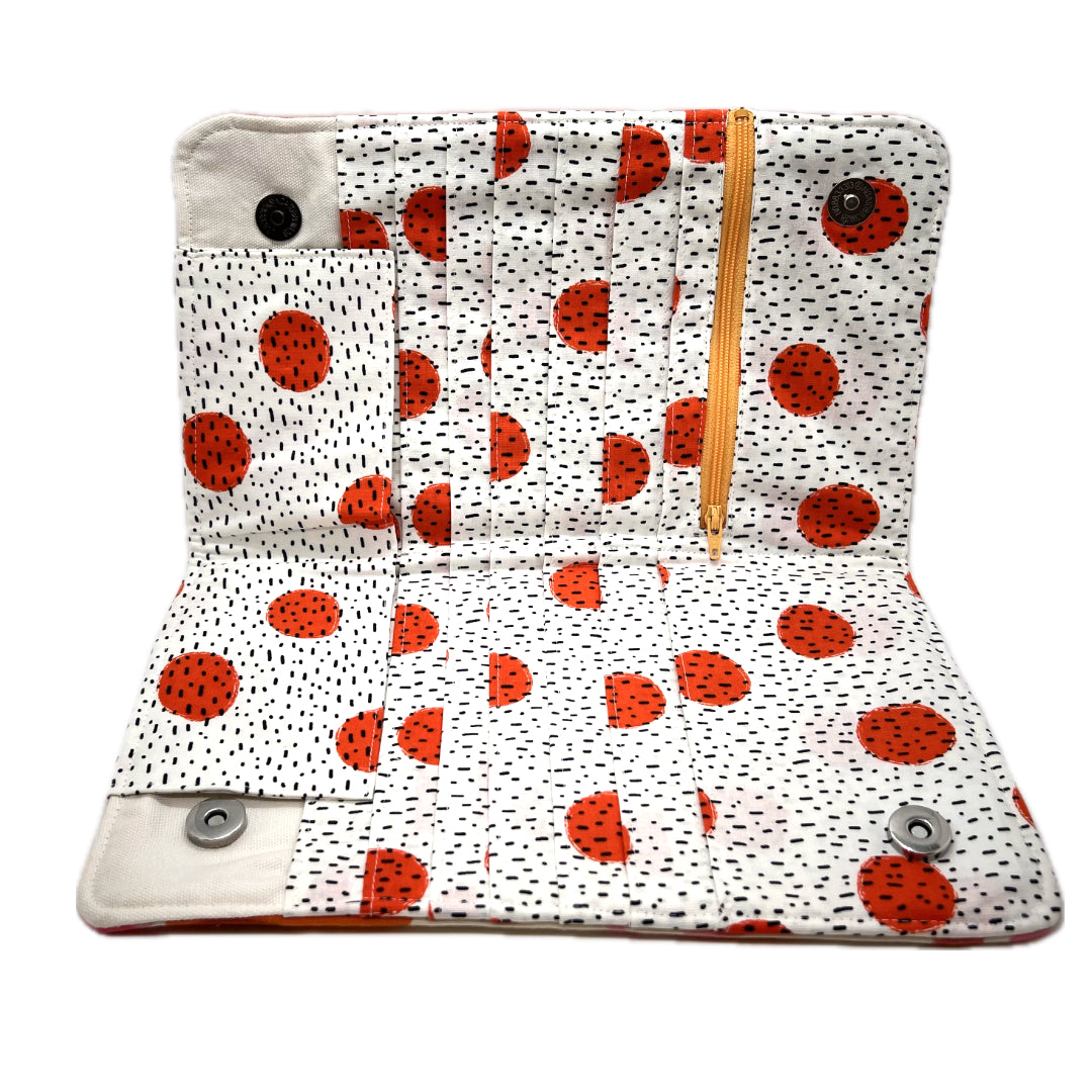 The inside of the needle organizer featuring its pockets in white fabric with bright orange circles and black speckles. The zipper on the notions pouch. 