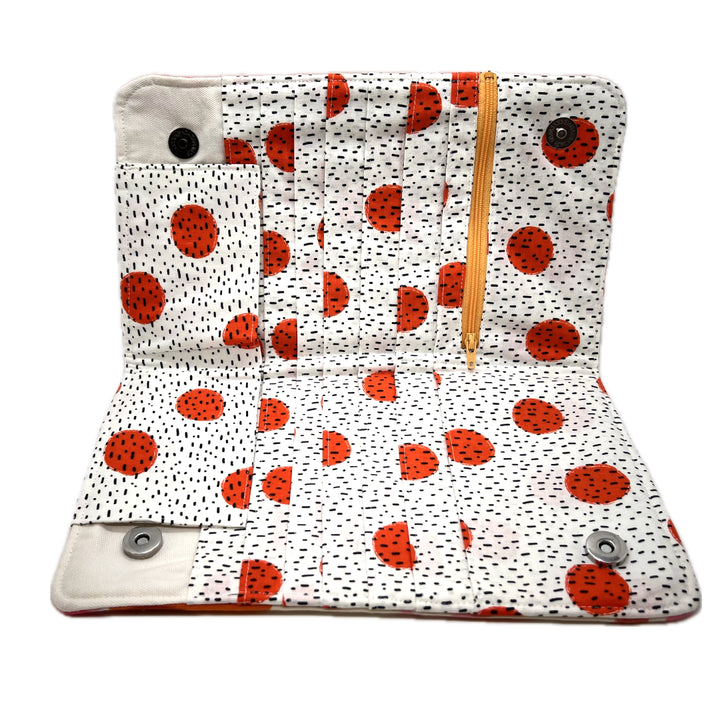 The inside of the needle organizer featuring its pockets in white fabric with bright orange circles and black speckles. The zipper on the notions pouch. 