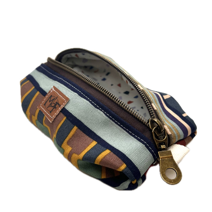 A zippered fabric pouch with a geometric pattern in navy blue, forrest green, orange with gray blue and brown stripes. The inside fabric has green, blue, gray and orange abstract confetti like shapes on a cream colored background. 