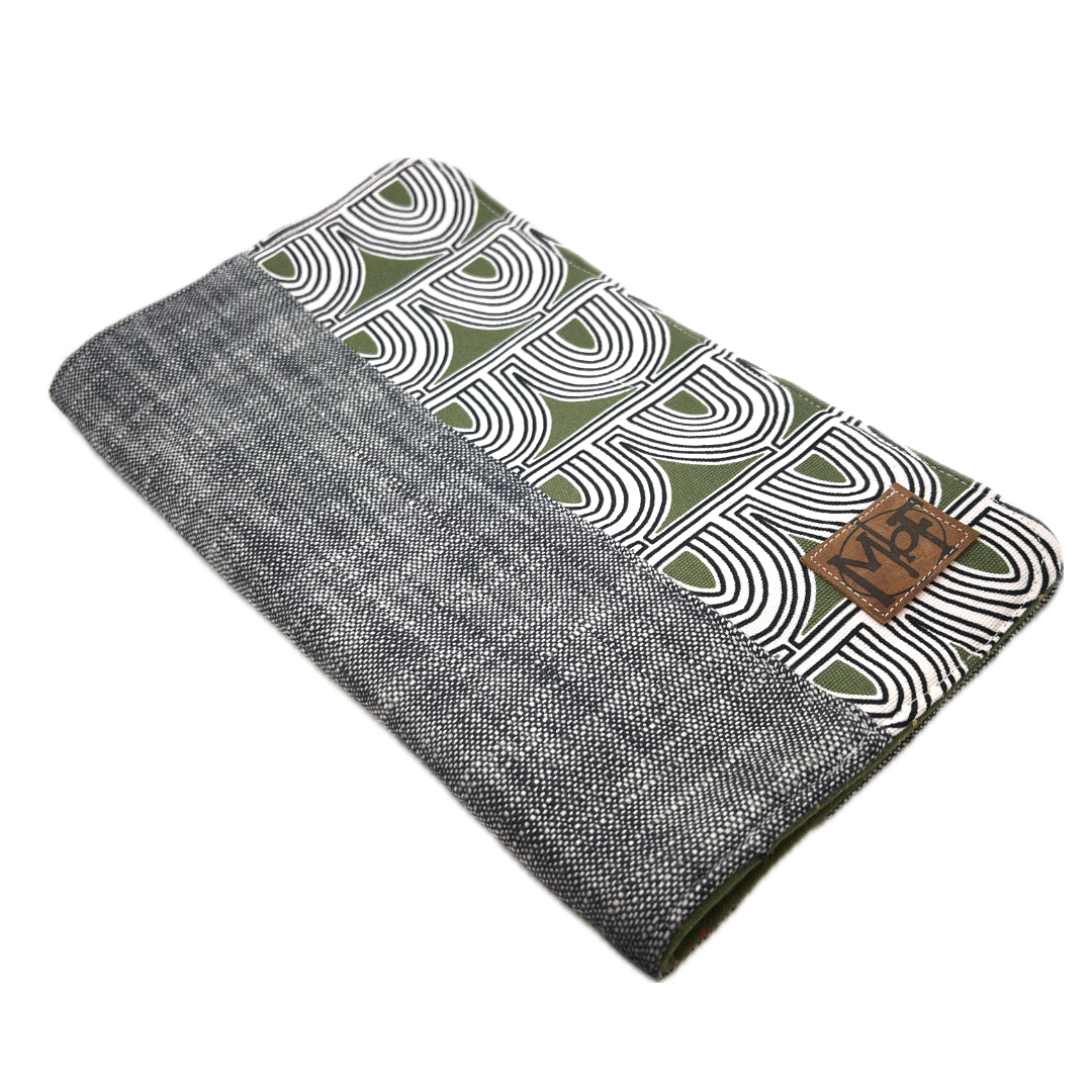 A closed handmade needle case. One side is a mossy green with concentric 1/2 circles in black on it, and the other side is a gray and black woven fabric. There's a leather patch that says MbF.