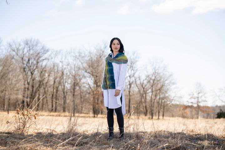 A fem person with shoulder length dark hair wearing the Elegante Wrap by Bobbi Intveld over a long white tunic and black leggings. She's standing in a field and the background is a fall scene with bare trees, and a blue/gray sky. 
