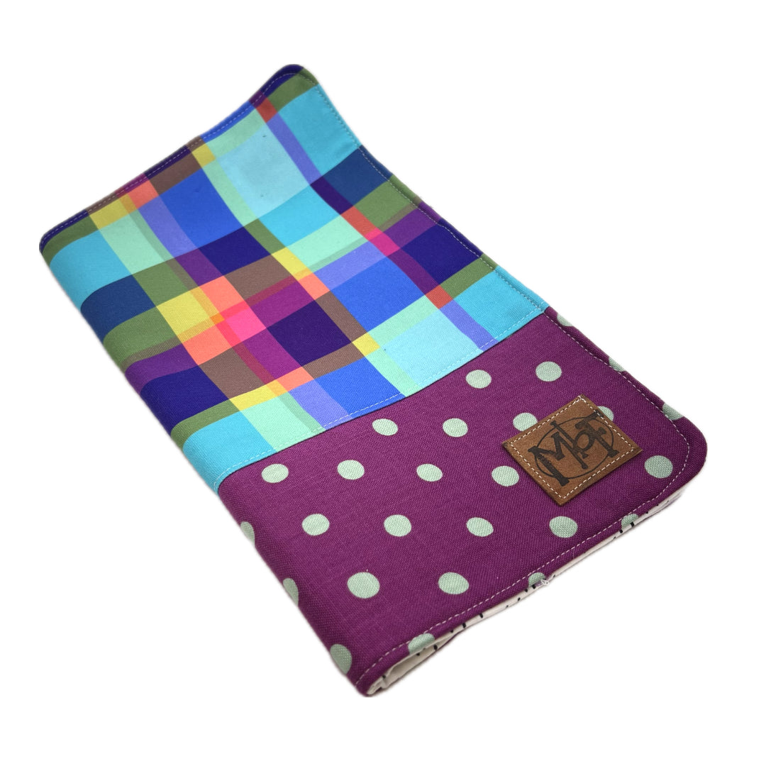 Front of a needle organizer case featuring a multicolor plaid pattern on the upper section and a purple polka-dot section on the lower section with a small brown leather patch that says MbF.