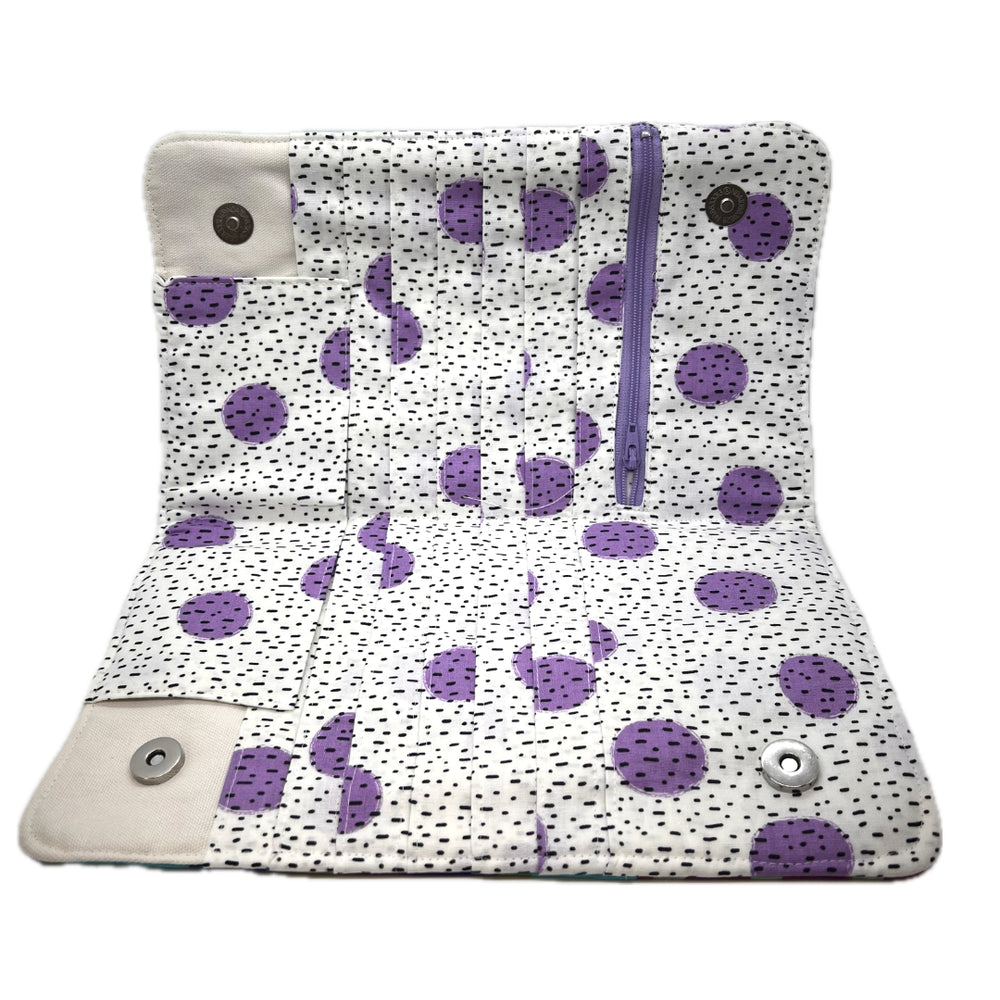 Inside of the needle case showing the pockets in a white fabric body with purple polka dots, small black speckles, a purple zipper.