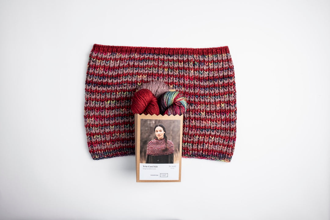 The Trio Cowl with the kid that creates it on top of it. There are 3 skeins of yarn, a deep red, a variated skein in jewel tones, and a mauve tweed skein. The cowl is in squishy soft ribs. 