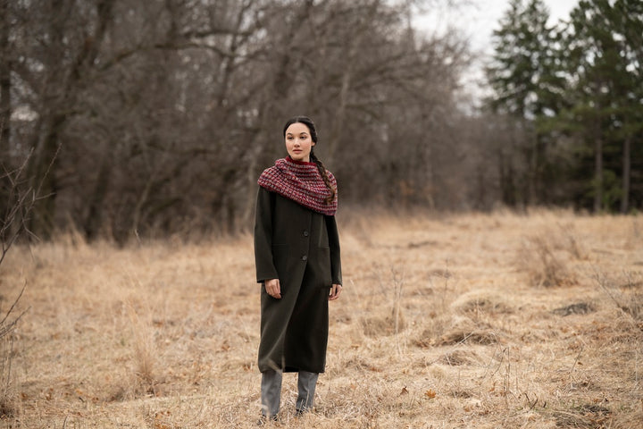 A fem person in an olive green coat with a warm cowl, predominantly in dark and muted reds. She's standing in a fall scene of prairie gone to seed, a stand of leafless trees, and some evergreens. 