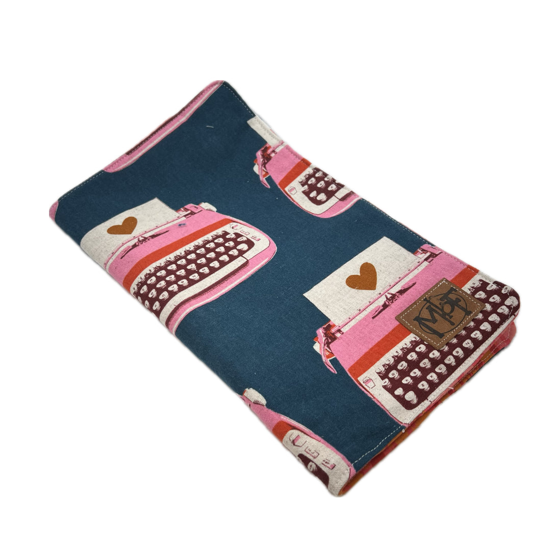 Front of knitting needle organizer featuring a repeating pattern of pink typewriters on a dark teal-blue background, with a brown leather MbF patch on the lower corner. 