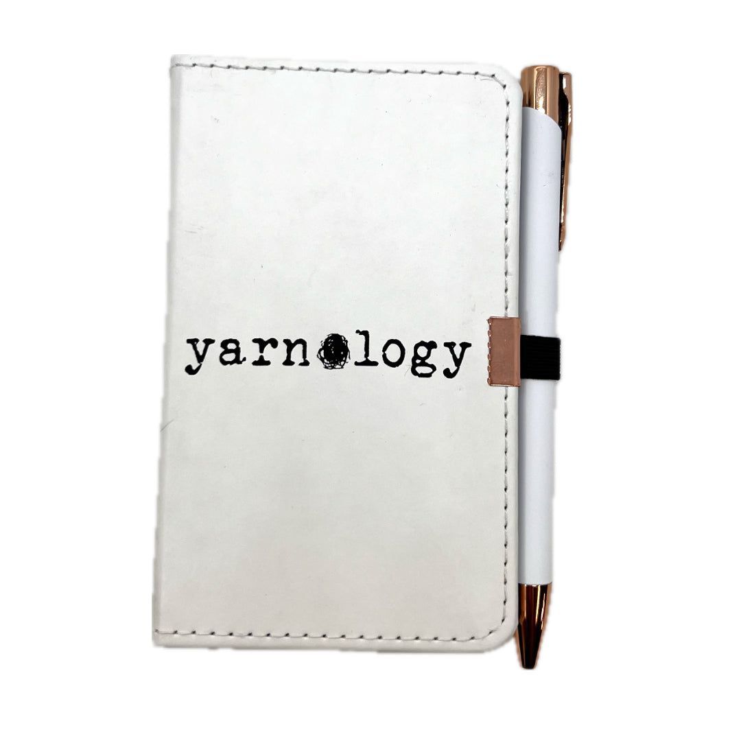 A white faux-leather pocket notebook with black stitched edges and the word 'yarnology' printed on the front, paired with a white pen that has rose gold accents and a black elastic band.