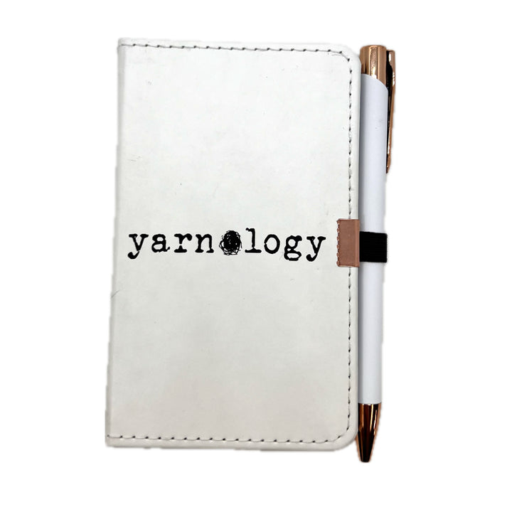 A white faux-leather pocket notebook with black stitched edges and the word 'yarnology' printed on the front, paired with a white pen that has rose gold accents and a black elastic band.